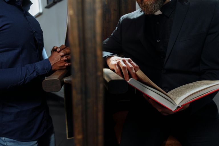 A man in a blue shirt with folded hands sits across from a priest reading a book during confession inside a church