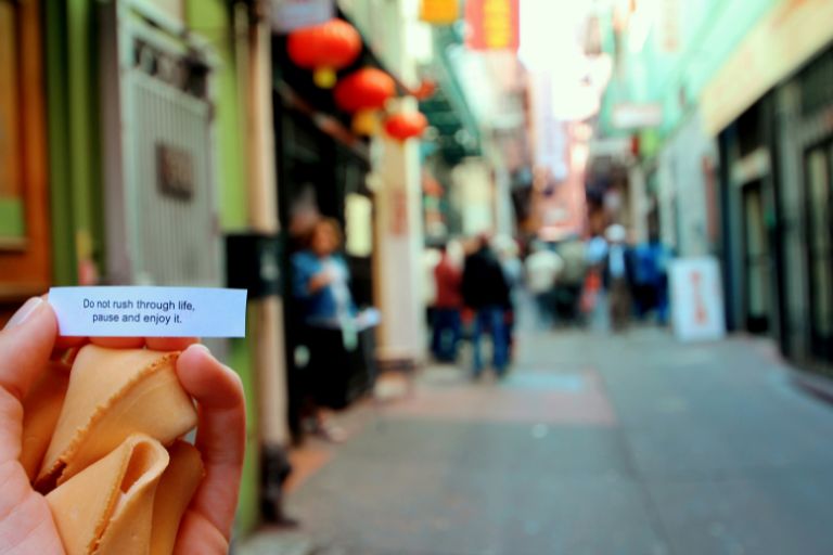 Hand holding fortune cookies and a message that reads “Do not rush through life, pause and enjoy it,” with a blurred city street and people in the background