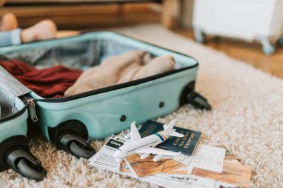 An open turquoise suitcase sits on a carpet with clothes inside, next to scattered travel documents, passports, and a small toy airplane