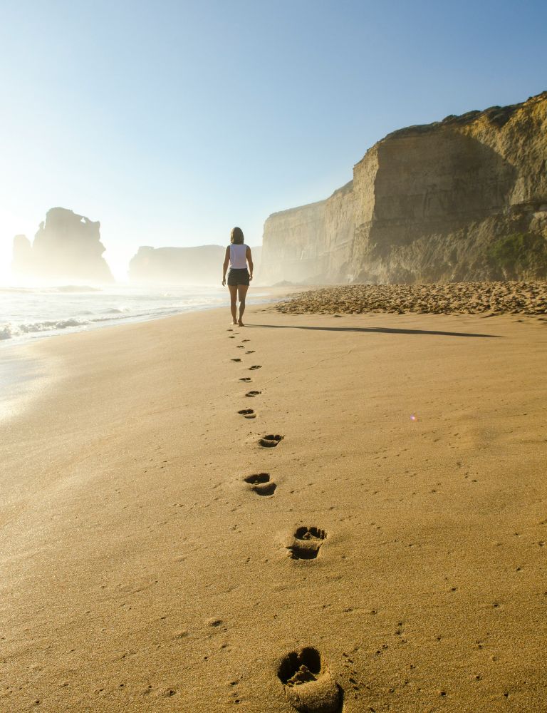 A woman walking alone on a sandy beach, leaving a trail of footprints behind her, with towering cliffs and rock formations in the background under a clear blue sky