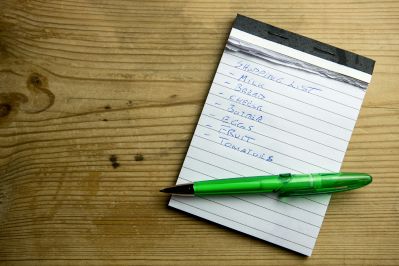 A handwritten grocery list on a notepad with a green pen placed on a wooden table