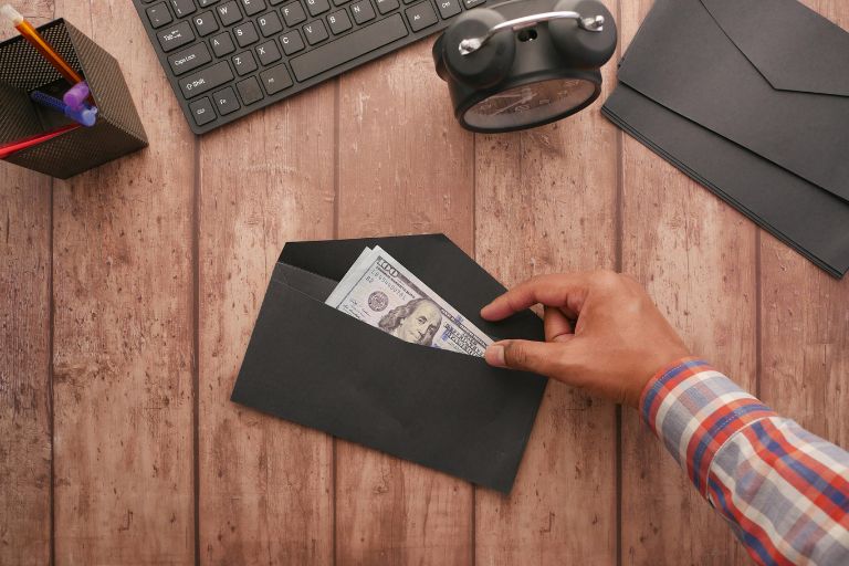 A person placing U.S. dollar bills into a black envelope on a wooden desk, with a keyboard, alarm clock, and office supplies nearby