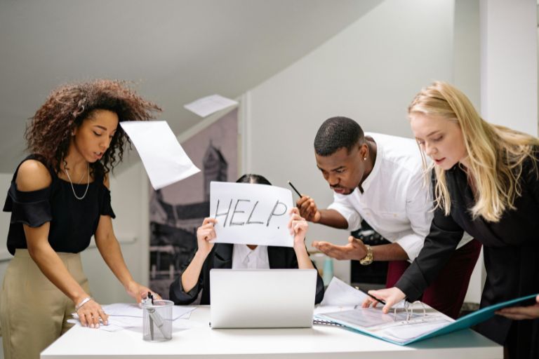A stressed office worker holding a 'HELP' sign at a chaotic desk, surrounded by coworkers and flying papers