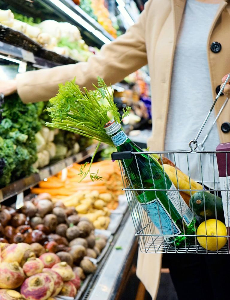 A woman wearing a beige coat shopping for vegetables with a handheld grocery basket full of fresh produce and bottled drinks
