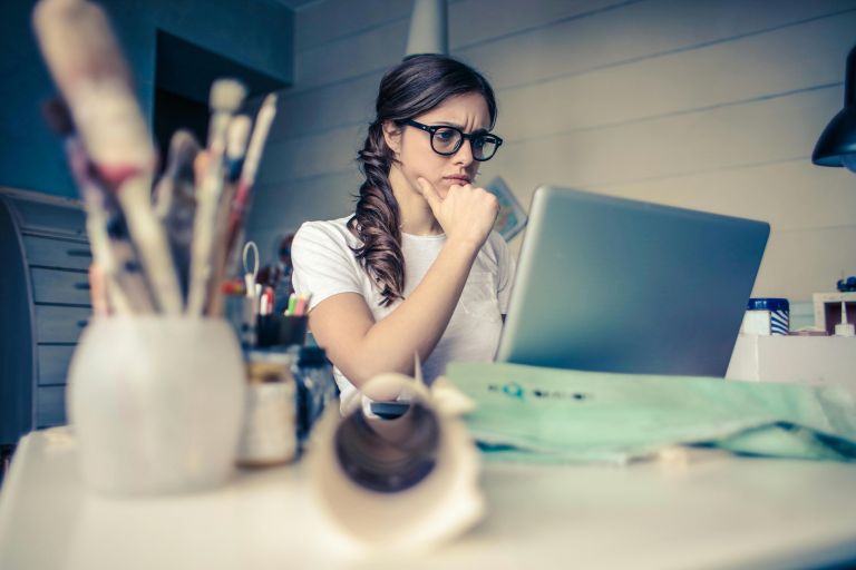 A thoughtful woman wearing glasses sits at a desk, looking at a laptop screen in a creative workspace