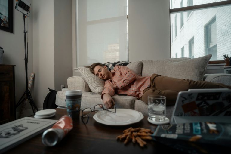 Young man lying on a couch surrounded by snacks, empty plates, and clutter—visibly drained, symbolizing burnout and loss of motivation