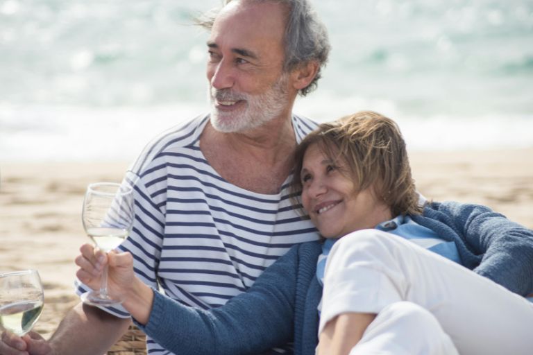 Smiling older couple sitting on the beach together, enjoying wine and a relaxing moment by the ocean