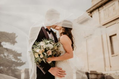 Bride and groom embracing intimately while bride holds white and green floral bouquet, with historic stone building in soft, dreamy lighting