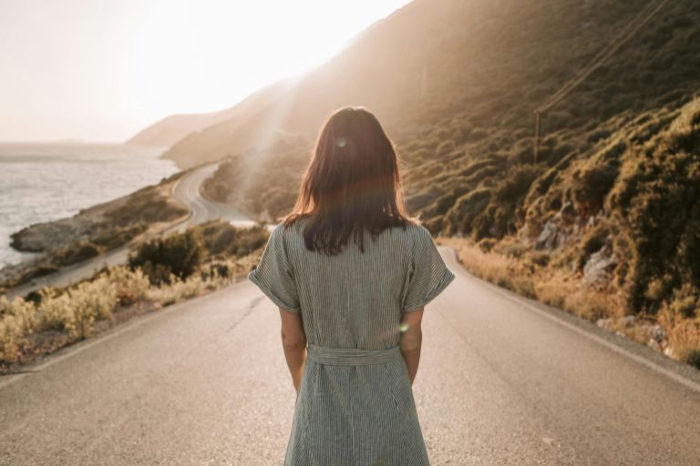 Woman in a striped dress standing on a winding coastal road at sunset, looking towards the horizon