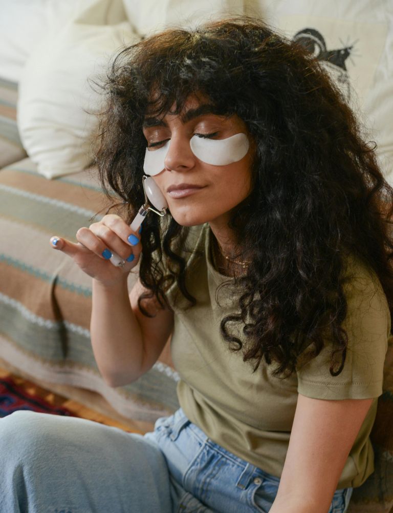Young woman with curly hair applying a face roller while wearing under-eye patches during a self-care routine at home