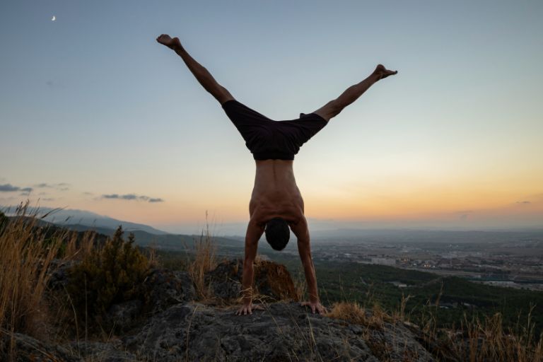 Person performing a handstand on a rocky mountain summit at sunset with a scenic valley landscape in the background