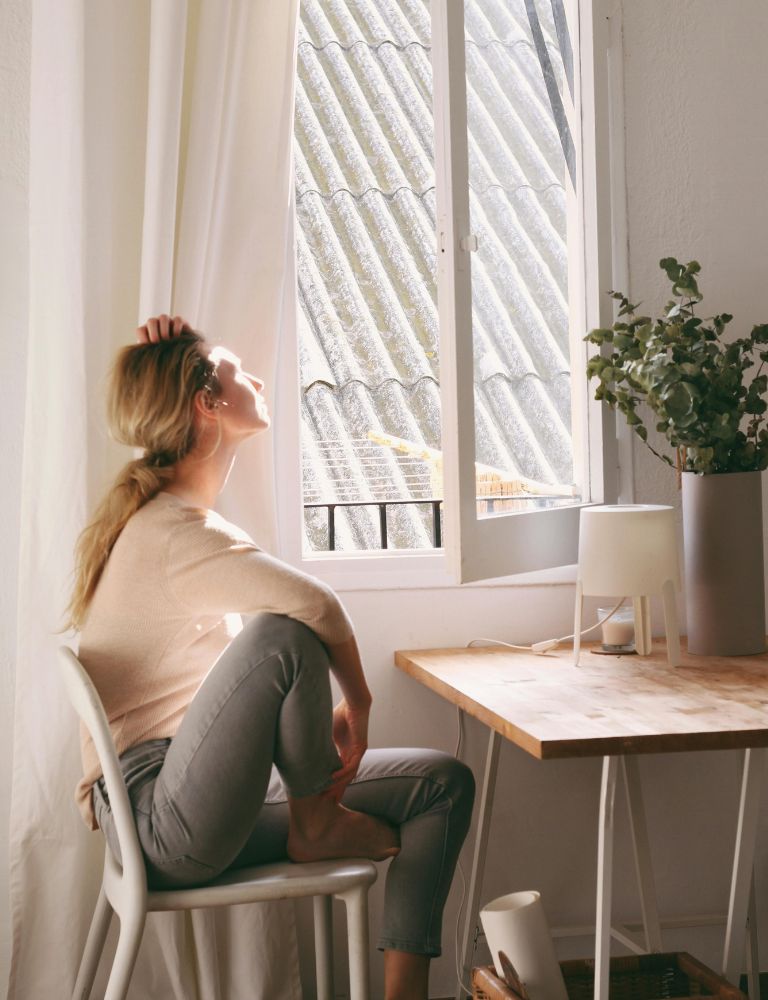 Woman sitting at desk by window with sunlight streaming in, head tilted back in relaxed pose