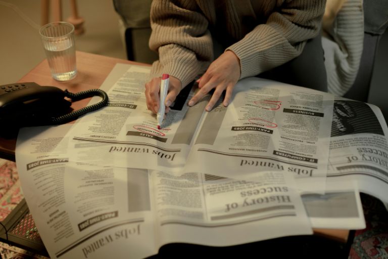 Person in green sweater circling job listings in newspaper want ads with red marker, representing traditional job search methods that often produce limited results